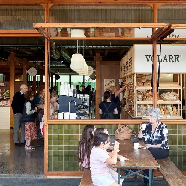 A busy market-style food hall or café frontage with an open timber-framed serving kiosk, green tiled counter, glass display shelving, pendant lights, exposed services and timber structure. Customers gather inside and sit at a rustic communal table in the foreground. The scene has a warm, lively hospitality atmosphere.