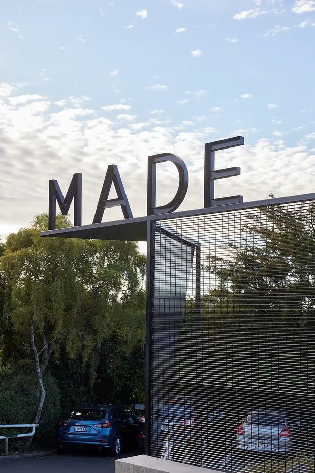 Exterior photograph of a modern parking or building entrance marked with large MADĒ lettering. The structure features a dark steel frame, extensive black wire-mesh screening, and a projecting flat canopy, with parked cars and mature trees visible around it.