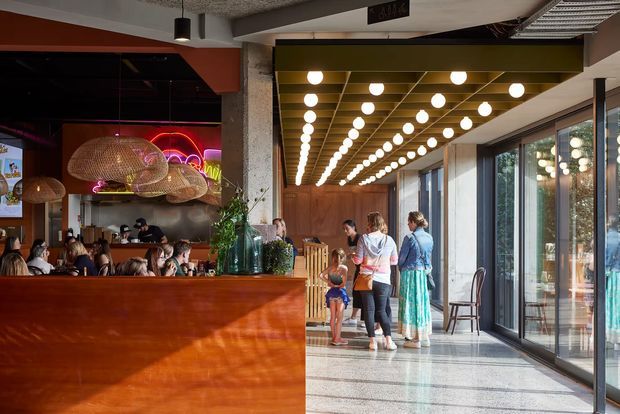 A lively contemporary restaurant interior with a prominent warm timber service counter and bar area, woven pendant lights, exposed concrete columns, a dramatic illuminated slatted ceiling, and full-height glazing along the right side. Diners and a small queue occupy the space, with polished flooring and strong indoor-outdoor visual connection.