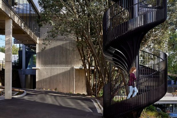 Exterior architectural photograph of a contemporary multi-level building with a dramatic sculptural black spiral staircase or ramp enclosed by vertical metal bars. Concrete columns, dark framed glazing, ribbed metal wall cladding, mature trees, and elevated walkways are visible. The composition emphasizes the expressive curved circulation element and modern facade.