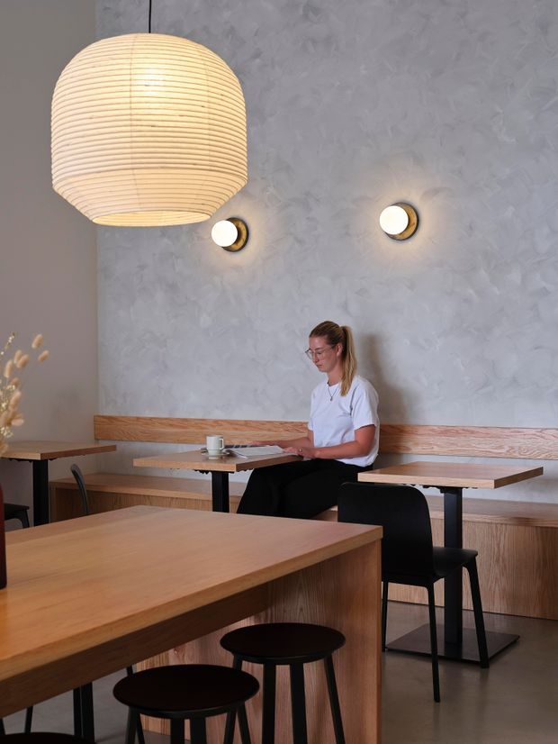 A softly lit contemporary café or hospitality interior with warm timber tables, built-in timber bench seating, black chairs and stools, and a textured pale grey plaster wall. A large woven pendant light and two small wall sconces provide warm ambient lighting. A woman is seated at a table with a laptop, reinforcing the café or workspace setting.