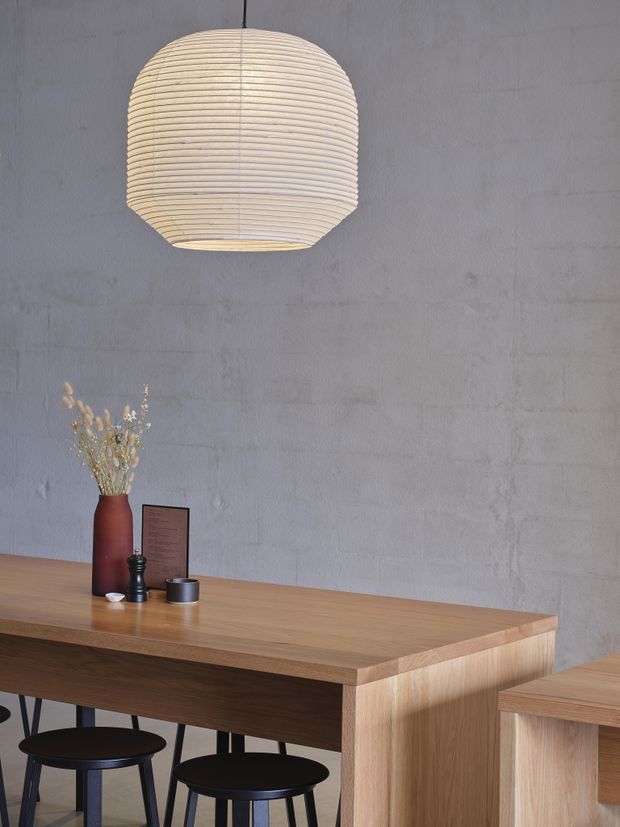 A minimalist dining or hospitality-style setting with a large woven pendant light above a long light timber table and black stools. The space has a raw grey concrete wall, simple black tabletop accessories, a terracotta vase with dried botanicals, and a timber side counter or cabinet in the foreground.