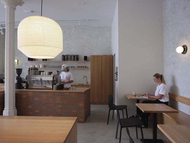 A refined café interior with a coffee service counter, espresso machine, open timber shelving, and wood-fronted cabinetry. A large ribbed pendant light hangs above the counter, while compact timber tables and black chairs line a built-in bench along a textured pale wall. Classical decorative columns contrast with the contemporary café fit-out.