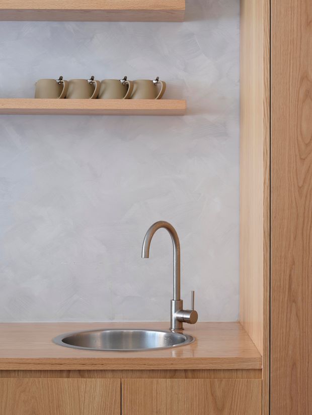 Close-up view of a compact kitchenette or kitchen cabinet run, featuring a stainless-steel round sink and curved mixer tap set into a light timber benchtop. Matching timber cabinetry and open shelves hold neutral ceramic mugs, while the wall has a soft textured pale grey finish. The composition is warm, minimal, and carefully detailed.