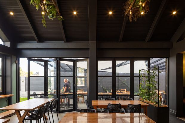 A dark, sophisticated dining and entertaining space with a high pitched ceiling, exposed dark timber beams, black-framed floor-to-ceiling glazing, and multiple timber dining tables. Black woven dining chairs surround the tables, while suspended greenery and warm pendant-style bulbs add an organic decorative layer. The glazing opens to a small courtyard or outdoor area, creating a strong indoor-outdoor connection.