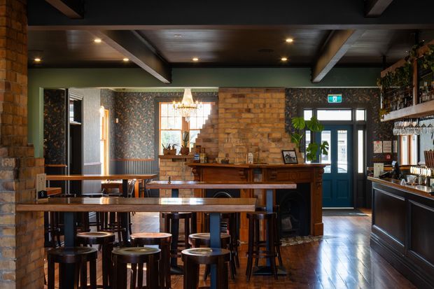 Interior view of a rustic, heritage-inspired bar or gastropub. The space features exposed brick walls and columns, dark timber ceiling beams and flooring, patterned feature wallpaper, long timber communal tables with stools, a substantial timber bar counter, pendant and recessed lighting, and a drinks service area with shelving and hanging glassware. A glazed entry door and windows bring in daylight at the rear.