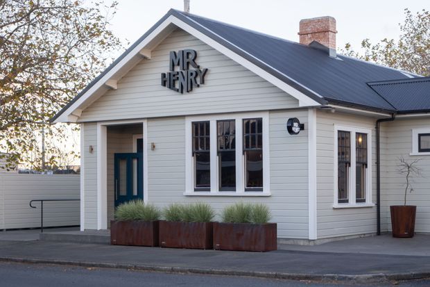 A small, single-storey gable-fronted building with pale horizontal weatherboard cladding, a dark metal pitched roof, white trim, and a brick chimney. The facade features a dark blue entry door, multi-pane timber-framed windows, planters with ornamental grasses, and signage reading “MR HENRY,” suggesting a cafe or small commercial venue. The building sits directly beside a paved street and driveway.