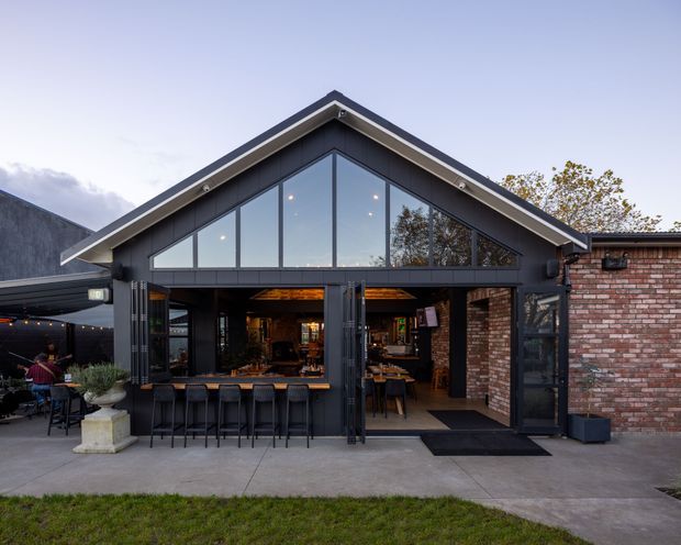 Exterior view of a contemporary gable-roof hospitality or commercial building with a dark metal-framed glazed facade, large triangular clerestory windows, exposed brick walls, and wide bi-fold or folding doors opening onto an outdoor seating area. Bar seating and tables are visible inside and outside, with warm interior lighting contrasting against the cool exterior.