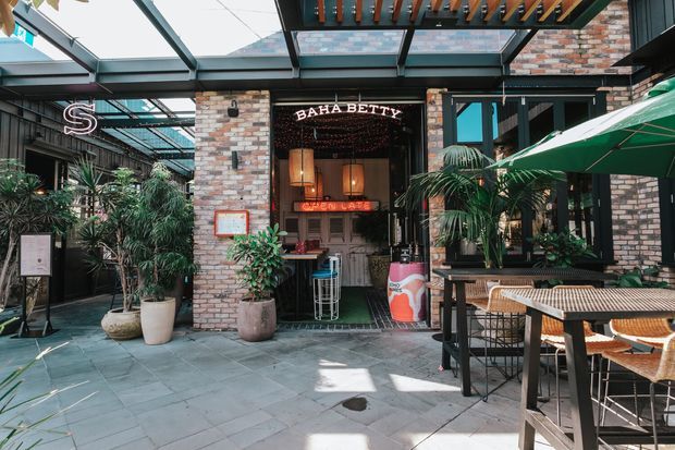 Exterior view of a stylish hospitality venue entrance and outdoor dining terrace. The facade combines exposed brick, black metal framing and extensive overhead glazing, with a covered pergola, tropical planting, green umbrellas, bistro tables and woven chairs. Signage identifies the venue as Baha Betty, with a neon-lit entry creating a lively urban restaurant atmosphere.