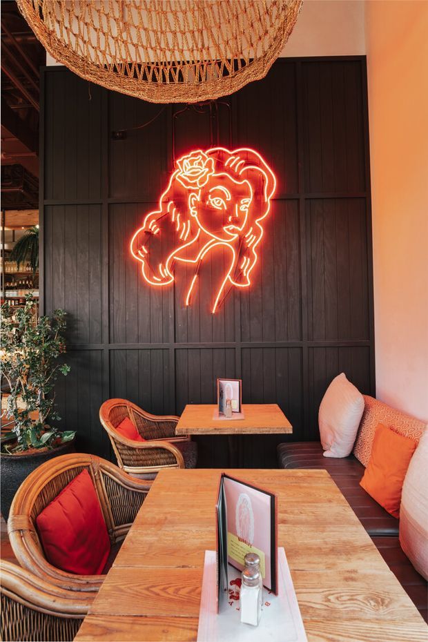 A warmly lit restaurant or hospitality interior with dark vertical timber wall panelling, woven rattan pendant lighting, timber dining tables, wicker chairs, upholstered bench seating, and a prominent red-orange neon line-art artwork. The space has an eclectic, intimate and slightly tropical character with abundant warm wood and orange accents.