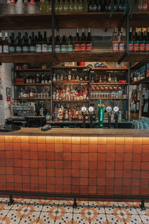 A richly stocked bar counter in a commercial hospitality interior. The foreground features a terracotta-orange square tiled bar front with a pale stone or composite countertop, while patterned floral encaustic-style floor tiles run along the base. Behind the counter are dark timber shelving and metal-framed storage filled with bottles, glassware, beer taps, and bar equipment. The space has a warm, vintage-inspired industrial character with dense product display and layered textures.