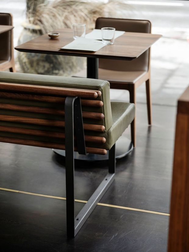 Close-up interior view of a refined dining or hospitality setting, featuring a green leather sling-back lounge chair with rounded timber slats and a black metal frame in the foreground. A dark timber dining table is set with glassware and tableware, with brown upholstered dining chairs behind it. The space has a dark smooth floor with a narrow brass inlay and soft natural light, creating a warm, understated mid-century atmosphere.