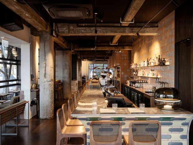 A long, narrow restaurant and bar interior with an industrial character. The space features exposed concrete columns, beams, ducts and dark ceilings, warm timber and metal detailing, a tiled bar counter with rounded multicolour tiles, open shelving filled with bottles and glassware, upholstered and woven bar seating, and large windows along one side.