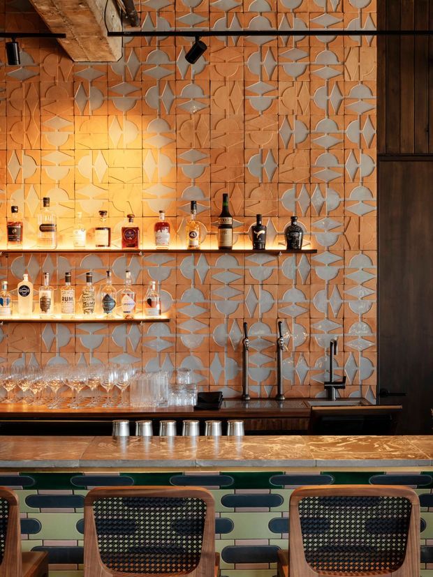 A warmly lit, stylised bar interior with a patterned terracotta and grey geometric tiled wall, backlit floating shelves displaying liquor bottles, a dark timber bar counter with brass-toned tapware, and a marble-effect benchtop. The foreground features upholstered bar stools with timber frames and woven backs, while black track lighting and dark timber joinery add an industrial, moody character.