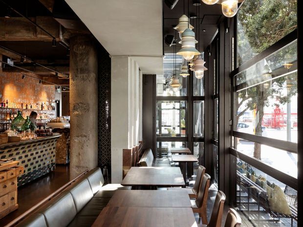 A stylish urban restaurant or bar interior with a long dining area beside floor-to-ceiling black-framed windows. Dark timber dining tables, leather banquette seating, and timber chairs line the glazed facade, while the adjacent bar features patterned tile cladding, exposed concrete columns, dark ceilings, and a copper-toned tiled backsplash. Suspended globe and layered pendant lights create a warm, eclectic atmosphere.