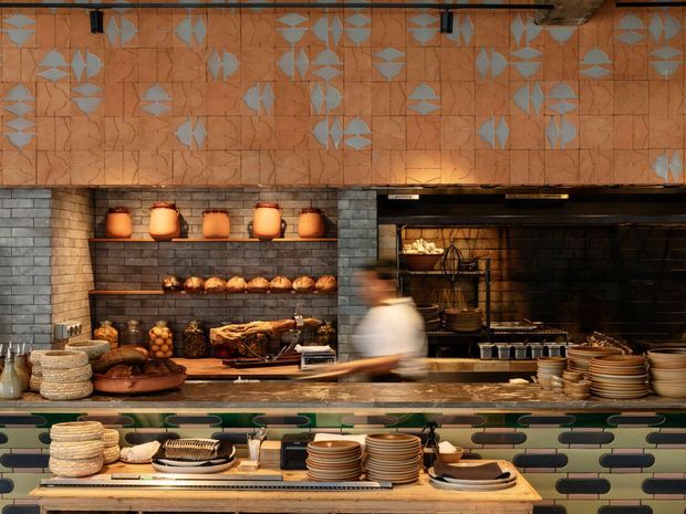 A warmly lit commercial kitchen or bakery counter with an open cooking and display area. The space features dark grey brick wall tiles, an orange patterned tile band across the upper wall, green and cream horizontal tiles at the service counter, timber shelving, ceramic plates and bowls, bread and produce displays, and a blurred staff member moving behind the counter. The aesthetic is richly textured, eclectic, and slightly industrial.