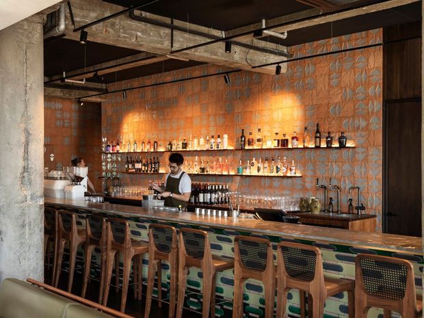 A warmly lit industrial-style bar interior with a long timber-fronted counter, rows of woven-back bar stools, open shelves displaying bottles and glassware, and a bartender workspace. The space features exposed concrete columns, visible services and ductwork, a dark ceiling, black metal framing, and a decorative orange and grey patterned tiled feature wall.