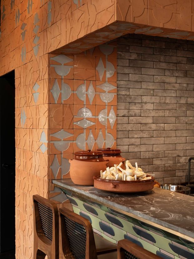 A richly detailed kitchen or bar counter clad in terracotta-toned decorative tiles with irregular geometric reliefs and metallic silver accents. The counter has a dark marbled stone benchtop and colourful patterned fixed cabinetry below, while the rear wall is finished in dark rectangular brick-like tiles. Warm lighting, timber-framed woven bar stools, ceramic vessels, and food create an earthy, eclectic atmosphere.