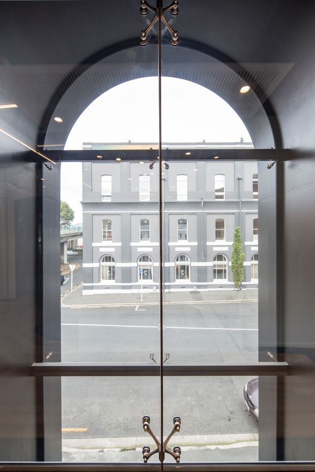 A tall, dramatic interior view looking through a large arched glazed opening toward a grey heritage-style building across the street. The foreground features dark charcoal walls, a central vertical mullion or seam, exposed metal hardware, recessed downlights, and a reflective glass partition or window system. The composition emphasizes the arched architectural opening and urban streetscape beyond.