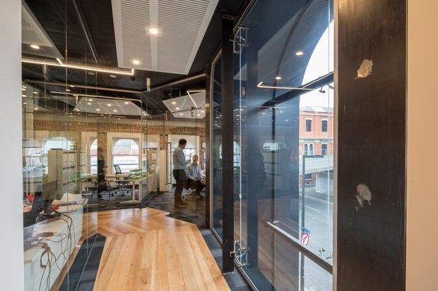 A contemporary commercial office interior viewed through glazed meeting-room partitions. The space features exposed dark ceilings, linear integrated lighting, acoustic ceiling panels, extensive glass walls and doors, timber flooring, and a large timber meeting table. Workstations and people are visible within the office, with an urban streetscape seen through the windows.