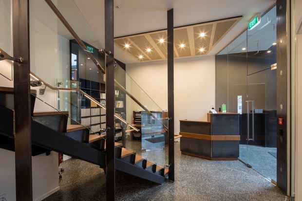 A contemporary commercial reception and circulation area viewed from the entry, featuring a black steel staircase with glass balustrades, a dark reception desk, polished speckled flooring, dark glazed walls, and a decorative perforated ceiling with recessed spotlights. Reflections in the glazing reveal additional office or lobby space.