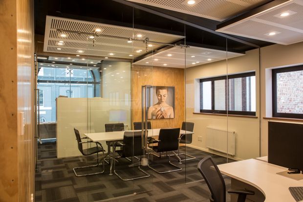 Contemporary commercial meeting room enclosed by full-height glass partitions, with a central conference table and black cantilever chairs. The room features warm timber wall panelling, dark carpet tile flooring, recessed downlights, and a slatted acoustic ceiling, with adjacent workspaces visible through the glazing.