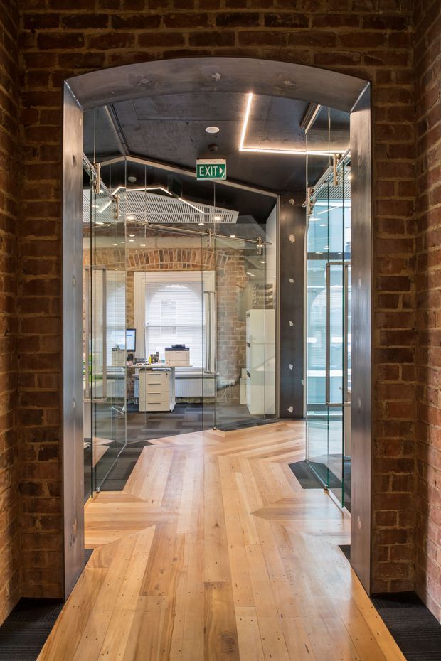 View through a large arched opening into a converted office interior. Exposed red brick walls surround a polished metal-framed glass entry, with warm timber parquet flooring, exposed concrete ceilings, black finishes, glass partitions, industrial-style metal railings, and visible workstations. Linear recessed lighting and an illuminated exit sign add contemporary commercial detailing.