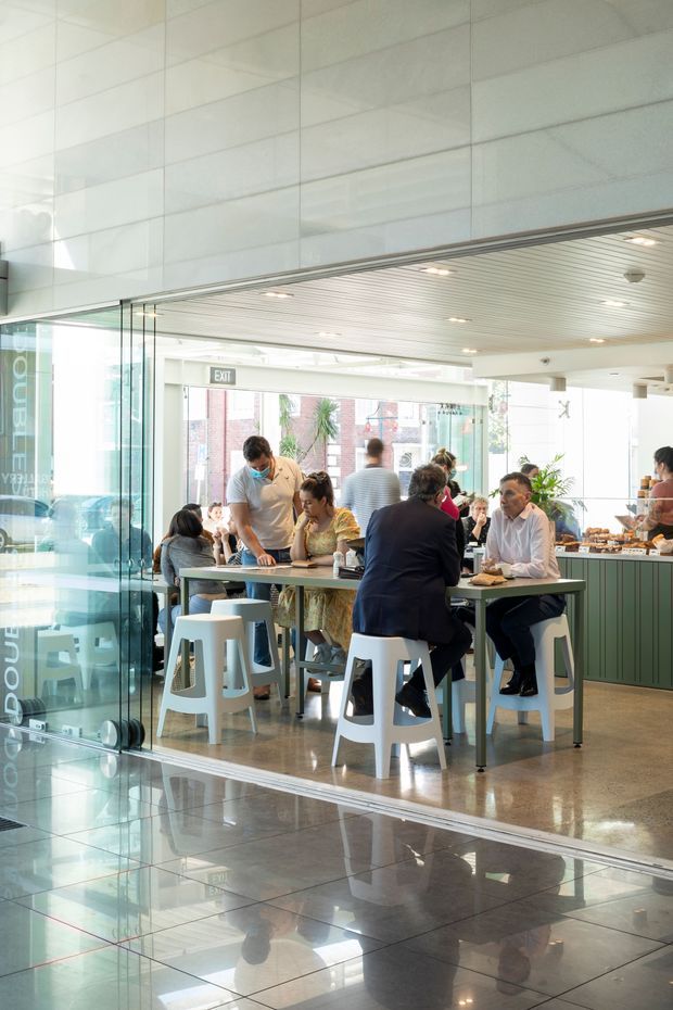 Bright contemporary commercial café or staff dining space viewed through a glazed frontage. Several people sit at communal tables and stools, with a service counter and food display at the rear. The space has extensive glass walls, pale tiled wall surfaces, a linear suspended ceiling, polished reflective flooring, and green fixed cabinetry. Reflections are visible in the foreground glazing and floor.