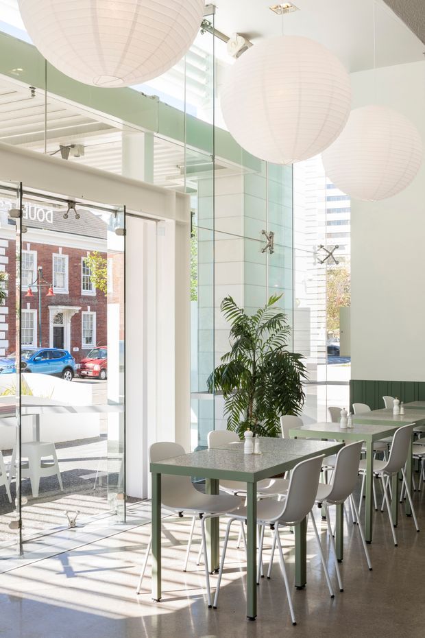 Bright, double-height café or casual dining interior with full-height glazed frontage, pale green and white finishes, rows of small green tables with white chairs, large spherical paper pendant lights, a tall indoor palm, and polished concrete flooring. Strong daylight enters through the glazing, with an urban streetscape visible outside.