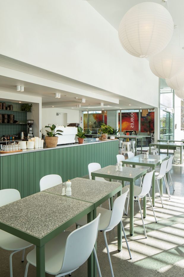 Bright contemporary café interior with a long green fluted service counter, terrazzo-style tabletops, white dining chairs and stools, indoor plants, and large white spherical pendant lights. Full-height glazing brings in abundant daylight, with additional seating and café areas visible beyond the counter.