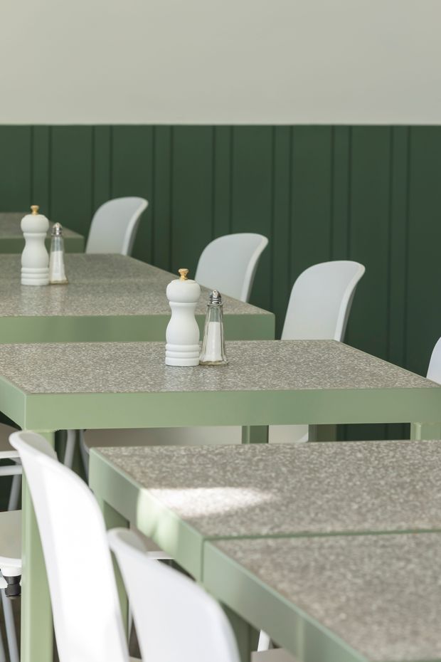 A close-up interior view of a contemporary dining or café setting with several pale sage-green rectangular tables topped with speckled stone-like surfaces. White molded chairs surround the tables, while white salt and pepper mills sit on the tabletops. The background features dark green vertical wall panelling, creating a calm, coordinated green-and-white palette with soft natural light.