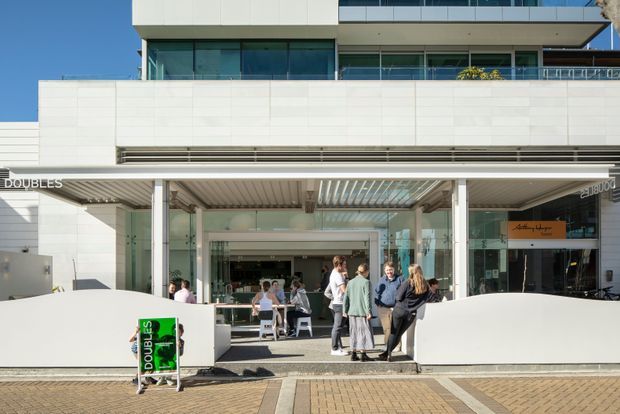 Exterior view of a contemporary multi-storey commercial building with a broad white tiled or panelled facade, extensive glass storefronts, upper-level glazing and a covered pedestrian entrance. People gather outside the open retail or hospitality frontage, with paved forecourt and horizontal metal canopy detailing.