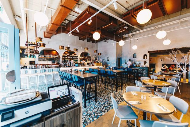 A spacious, brightly lit contemporary restaurant and bar interior with a long turquoise-accented bar, high communal tables, round dining tables, and a patterned tile floor. The space features exposed dark ceiling services and ductwork, white-painted walls, globe pendant lights, arched bottle-display niches, and warm timber and brass-toned details. A visible service counter and point-of-sale area occupy the foreground.