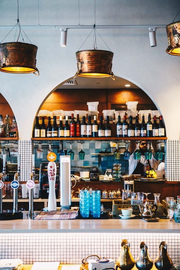 A stylish bar and hospitality interior viewed from the customer side of the counter. The space features a tiled bar front and backsplash, dark timber shelving and cabinetry, arched wall niches, extensive wine and spirits displays, hanging copper-toned pendant lights, glassware, beer taps, and a polished service counter. The overall atmosphere is warm, vibrant, and eclectic.