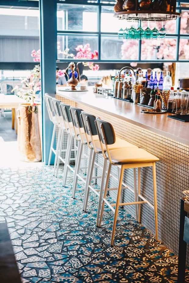 A stylish café or bar interior featuring a long tiled counter with yellow upholstered bar stools, a patterned encaustic-style tile floor, dark teal structural framing, large industrial-style windows, and a suspended overhead glassware rack. The counter is arranged with cocktail and beverage service items, while bright floral elements and seating are visible in the background.