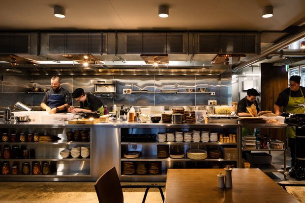 A warmly lit commercial open kitchen viewed from the dining area, with chefs working at stainless-steel preparation benches, extensive overhead ventilation and shelving, and open shelves displaying stacks of plates, bowls, and kitchen supplies. A wooden dining table occupies the foreground.