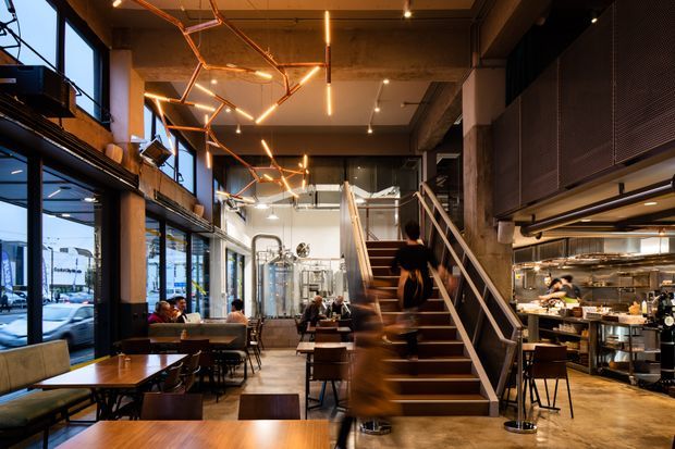 A contemporary double-height restaurant interior with industrial detailing, warm timber dining tables, booth seating, exposed concrete and metal elements, large street-facing windows, and an open commercial kitchen. A prominent central staircase leads to an upper level, while geometric linear pendant lighting creates a warm focal feature.