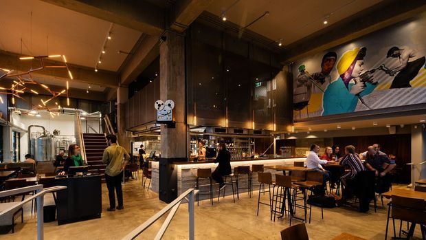 A spacious, double-height contemporary restaurant or bar interior with an open kitchen and long illuminated counter, surrounded by high tables and stools. The space features dark timber wall cladding, exposed structural beams, polished concrete flooring, industrial-style track and pendant lighting, and a large colourful mural above the dining area. A stairway and mezzanine-like upper level are visible at the rear.