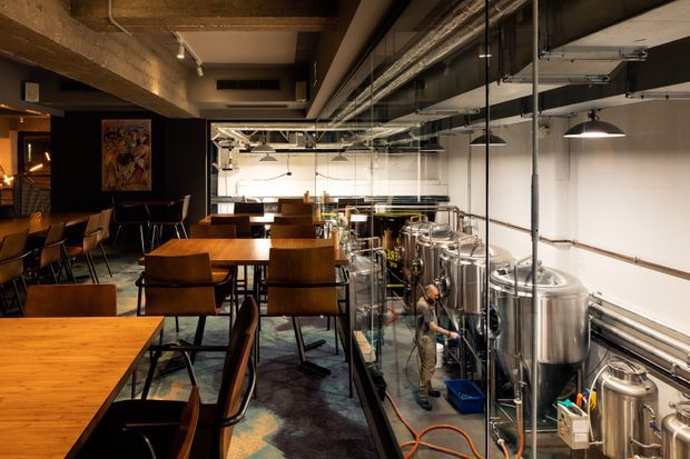 Industrial-style brewery taproom or dining space with timber tables and leather-upholstered chairs on the left, separated from visible stainless-steel brewing equipment by a glazed partition on the right. The room has exposed concrete beams, exposed services and ductwork, track and pendant lighting, dark flooring, and a warm, moody hospitality atmosphere.