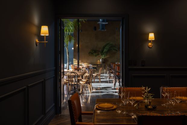 A moody, refined dining interior viewed through a dark paneled threshold, with warm wall sconces, a timber dining table, upholstered chairs, glassware, and a brighter adjoining restaurant-style dining area beyond. The space features dark wall panelling, polished timber furnishings, indoor planting, and strong natural light entering through glazing.