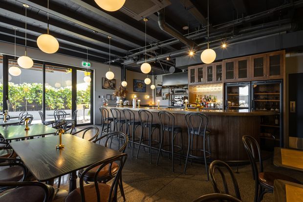 A warmly lit industrial-style bar and dining venue with dark timber tables, black bentwood chairs, a long timber-fronted bar, glass-front cabinetry, exposed black-painted ceiling services and ductwork, and numerous spherical pendant lights. Large glazed doors open toward greenery outside.