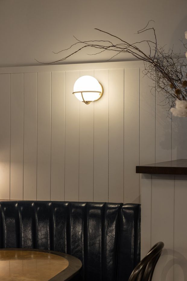 A moody dining nook with a dark leather channel-tufted banquette, rounded wooden dining table, and a partially visible chair. The room features warm wall lighting, cream vertical wall panelling, and a dark projecting shelf or counter. Decorative bare branches extend across the upper corner.