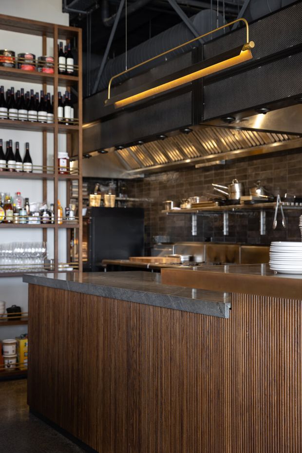 A sophisticated commercial hospitality kitchen and service counter, viewed from the customer side. The foreground counter has dark stone edging and vertically fluted dark timber cladding, with stainless-steel work surfaces behind it. Open timber shelving displays wine bottles, glassware, and supplies. The rear kitchen features dark brown tiled walls, a large stainless-steel extraction canopy, exposed black ceiling services, and a linear brass-gold pendant light.