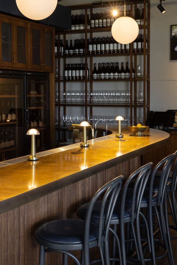 A warmly lit bar interior featuring a long polished amber-toned counter with integrated beer taps and small lamps, dark fluted timber bar frontage, curved black bar stools, and extensive open shelving displaying wine bottles and glassware. Dark timber cabinetry, black accents, pendant globes, and track lighting create an intimate, refined hospitality atmosphere.