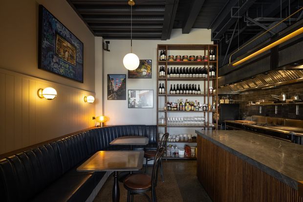 A moody industrial-style restaurant or bar interior with a long black leather banquette, small dining tables, timber wall panelling, framed artwork, warm globe wall and pendant lights, a tall open shelving unit stocked with bottles and glassware, and a large stone-topped bar or service counter beside an open commercial kitchen. The ceiling exposes dark structural beams and services, creating a refined hospitality setting.