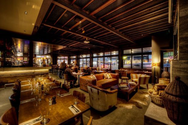 A warmly lit, rustic-industrial restaurant or hospitality interior with an open dining and bar area. The space features exposed dark timber ceiling beams and structure, large black-framed windows, a long bar counter, leather and upholstered lounge seating, timber dining tables, and woven decorative baskets. The atmosphere is intimate and richly layered, with amber lighting and dark natural materials.