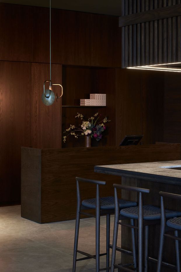A moody, refined kitchen and bar area with extensive dark timber wall panelling and cabinetry, a long island with a pale stone benchtop, and dark bar stools. A built-in timber reception-style cabinet includes open shelving with books and flowers, while a sculptural pendant and linear ceiling lighting provide warm illumination.
