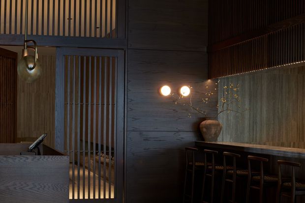 Moody, refined interior with dark stained timber wall panelling and vertical timber screens. A sculptural pendant hangs above a stone or concrete counter in the foreground, while a long built-in bar or dining counter with stools runs along the right wall beneath warm globe wall lights. The space has a high ceiling and strong linear detailing, with a restrained dark natural-material palette.