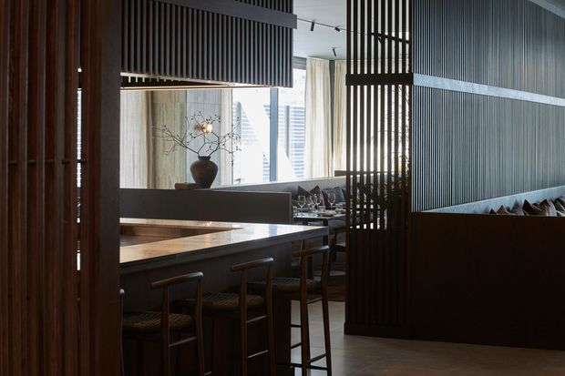 A refined contemporary dining and lounge interior with dark timber slatted screens creating partial partitions. A raised dark island or counter with bar stools occupies the foreground, while a dining table, upholstered seating, decorative pottery, and city views through full-height windows are visible beyond. The palette is dark timber, charcoal, warm neutral stone, and soft beige textiles, with layered linear lighting and a calm, sophisticated atmosphere.