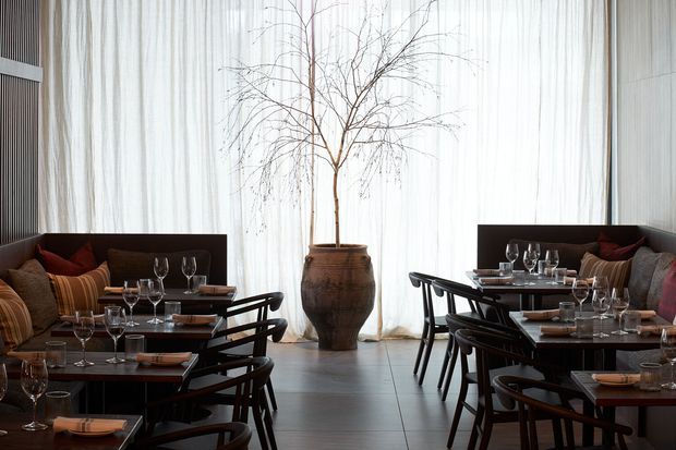 A refined contemporary dining room or restaurant interior with two rows of dark timber dining tables and chairs, upholstered banquette seating, table settings, and wine glasses. Full-height sheer curtains diffuse abundant daylight, while a large sculptural planter with a bare branch arrangement forms a central focal point. The palette is dark brown, black, warm timber, and muted red accents, creating an elegant atmospheric setting.